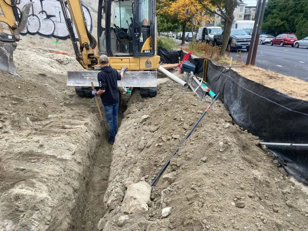 Excavator working on trench for underground utilities installation, with construction worker managing the site in Jacksonville area.