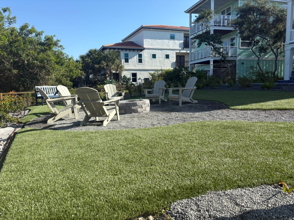 Outdoor seating area with white Adirondack chairs around a stone fire pit, surrounded by artificial grass and gravel, highlighting hardscaping features suitable for enhancing outdoor spaces in Jacksonville.