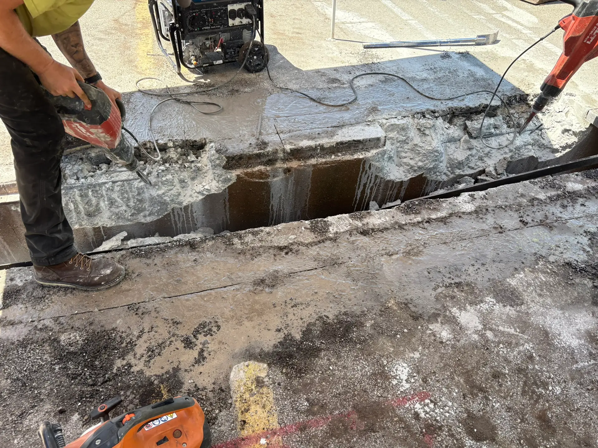 Worker using a jackhammer to repair a trench drain, with exposed concrete and water runoff, showcasing drainage solutions for property maintenance.